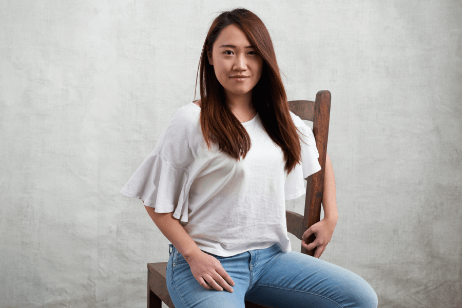 A young woman in a white shirt sits on a wooden chair facing the camera