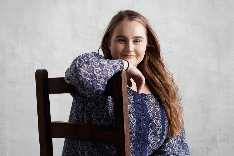 A young woman in a blue shirt sits on a wooden chair facing the camera